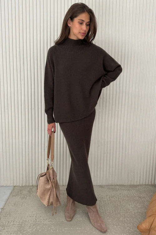 Woman wearing a dark brown outfit with a beige handbag against a light background