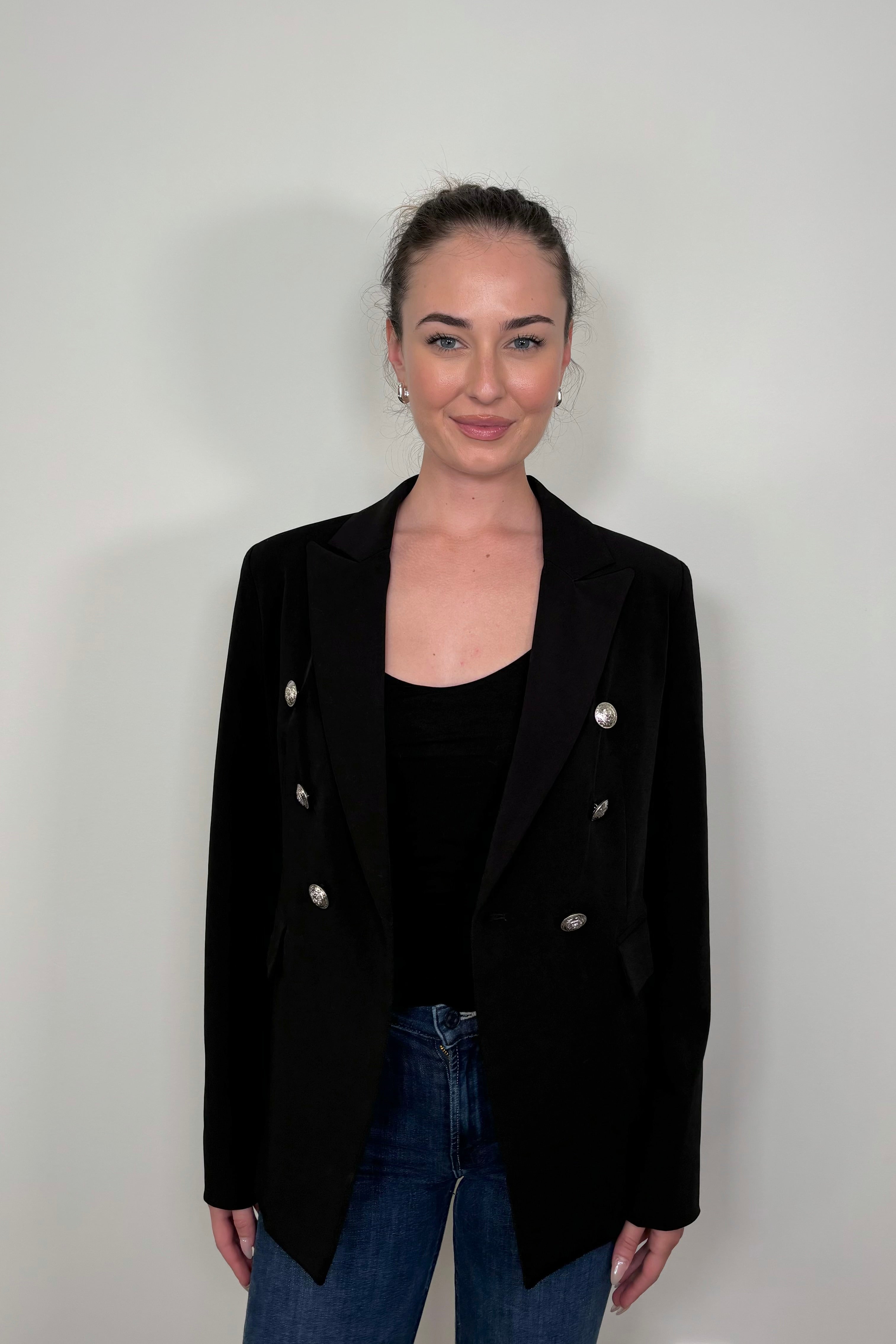 Woman wearing a black blazer with silver buttons on a plain background