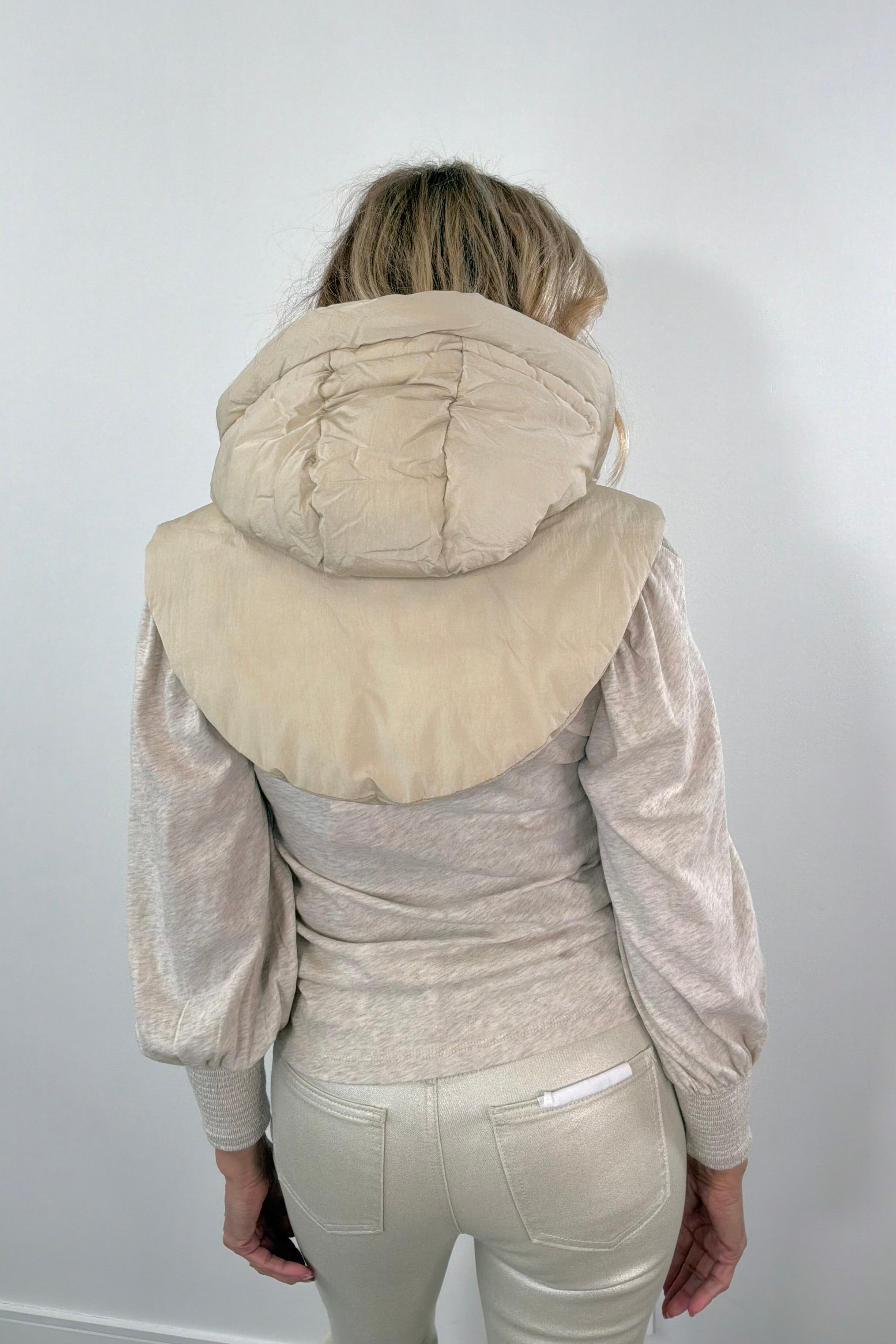 Woman wearing a beige puffer hood over a light-colored outfit against a plain background