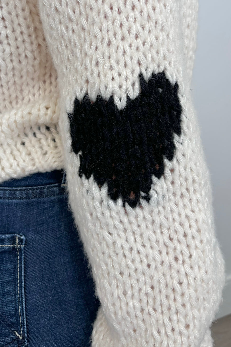 Close-up view of a person wearing a cream-colored sweater with a black heart design and whip-stitch detailing on the collar
