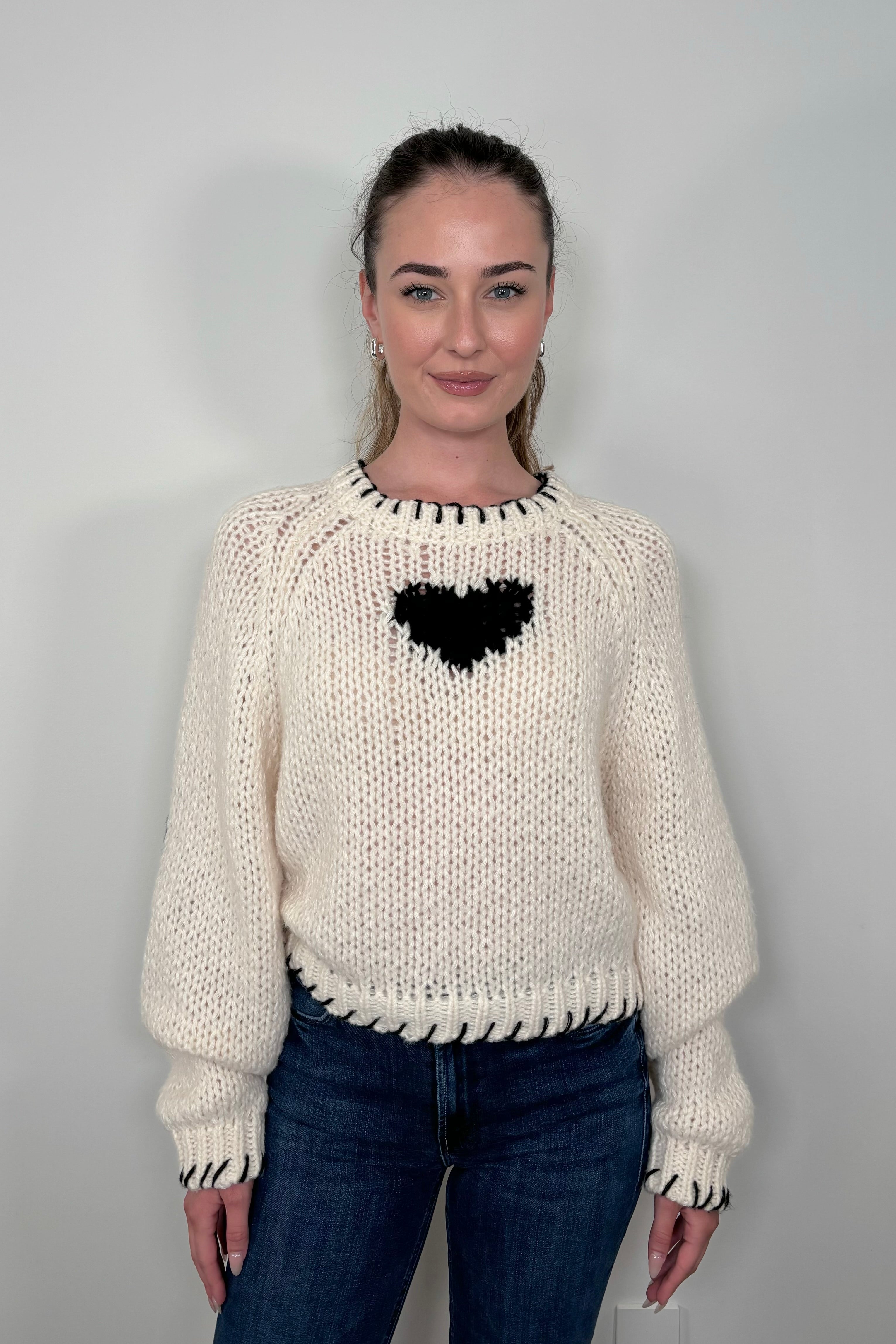 Woman wearing a cream knitted sweater with a black heart design against a plain background