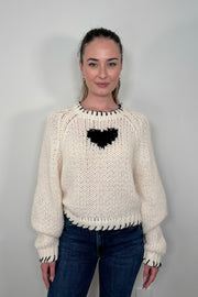 Woman wearing a cream knitted sweater with a black heart design against a plain background