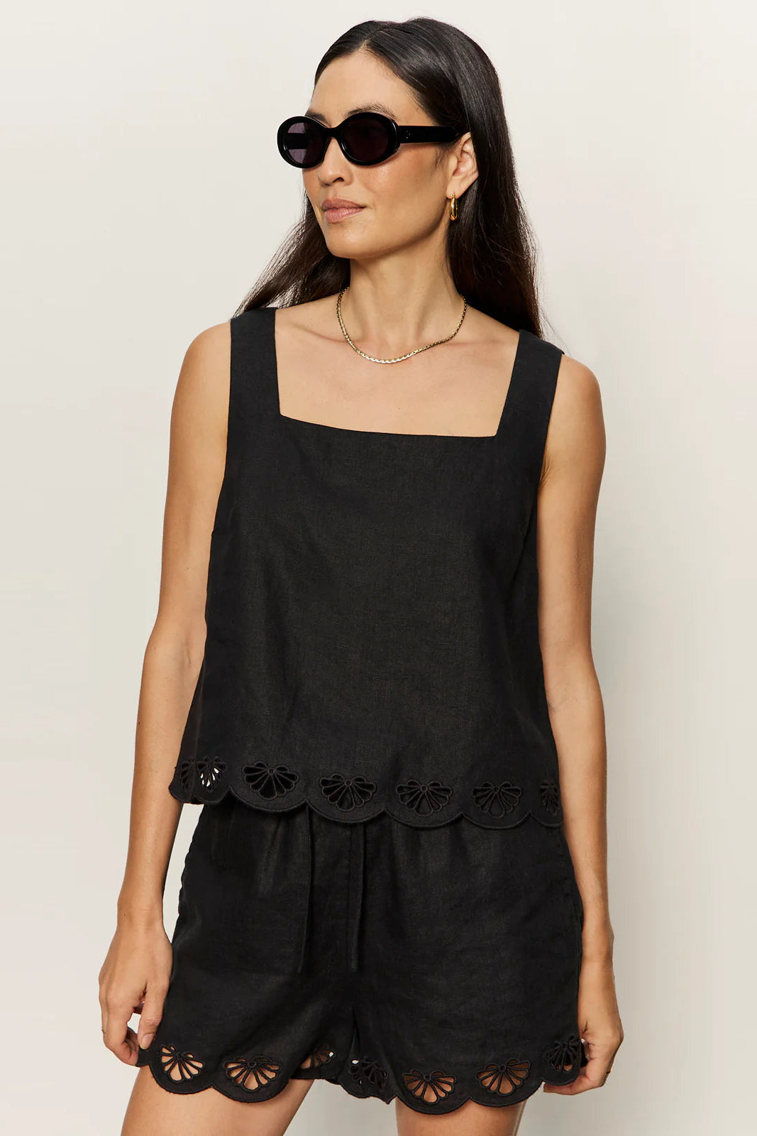 woman wearing a black sleeveless top with wide straps and scalloped hem detailing paired with matching shorts, front