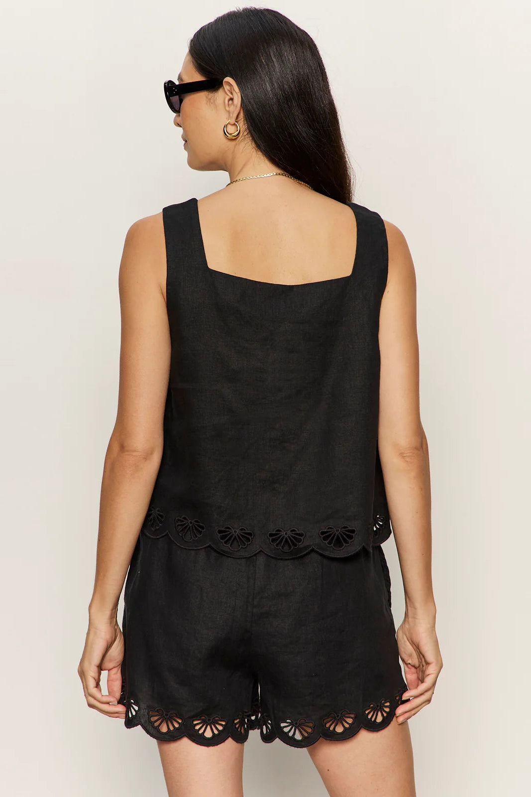 woman wearing a black sleeveless top with wide straps and scalloped hem detailing paired with matching shorts, back