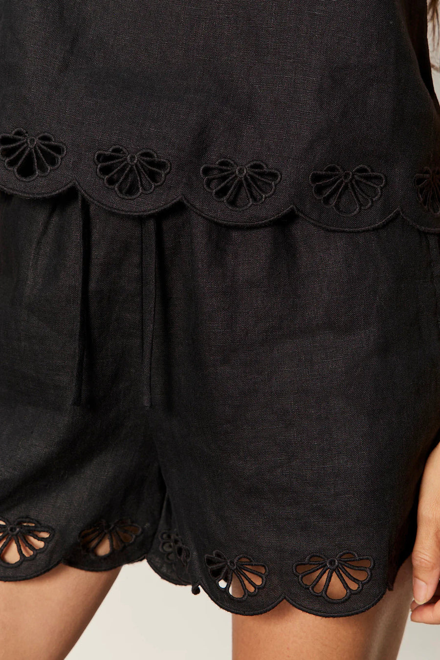 woman wearing a black sleeveless top with wide straps and scalloped hem detailing paired with matching shorts, detail