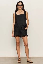 woman wearing a black sleeveless top with wide straps and scalloped hem detailing paired with matching shorts, front