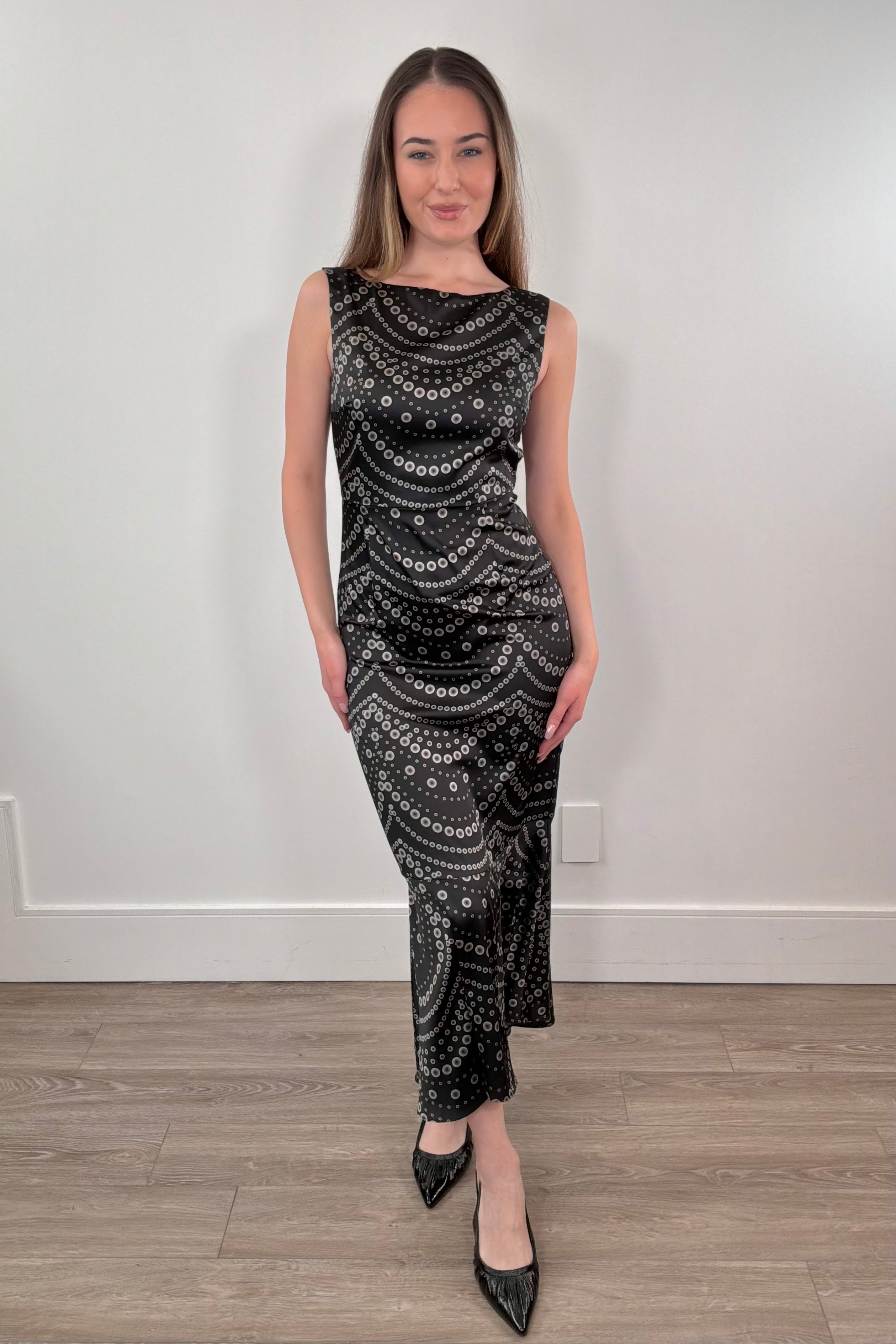 Woman wearing a black patterned dress standing against a white wall.