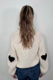 Back view of a person wearing a cream-colored sweater with a black heart design and whip-stitch detailing on the collar