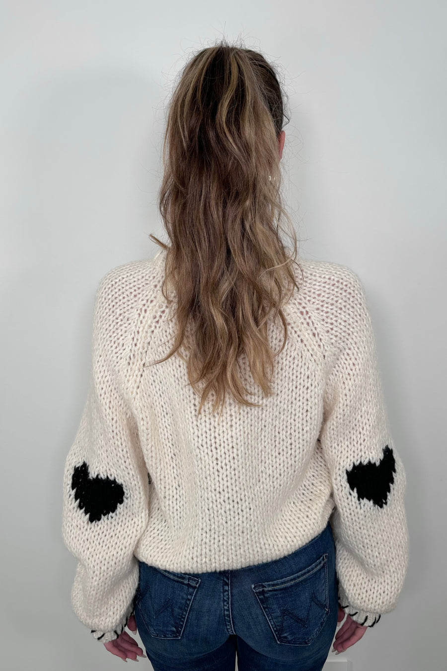 Back view of a person wearing a cream-colored sweater with a black heart design and whip-stitch detailing on the collar