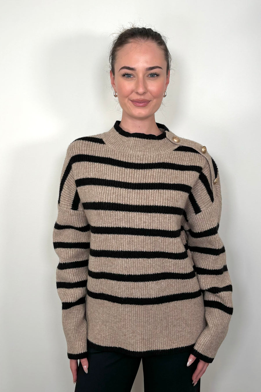 Front view of a woman wearing a walnut coloured sweater with stripes and buttons on the left shoulder