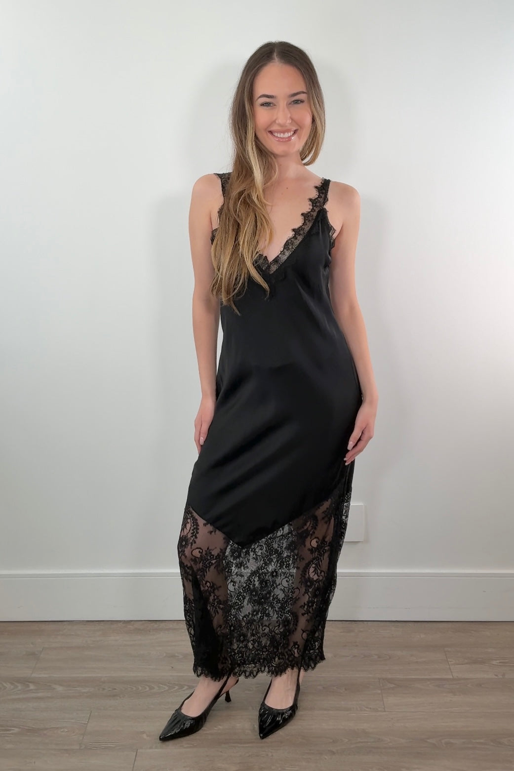 Woman wearing a black satin slip dress with lace details against a white wall.