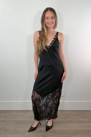 Woman wearing a black satin slip dress with lace details against a white wall.
