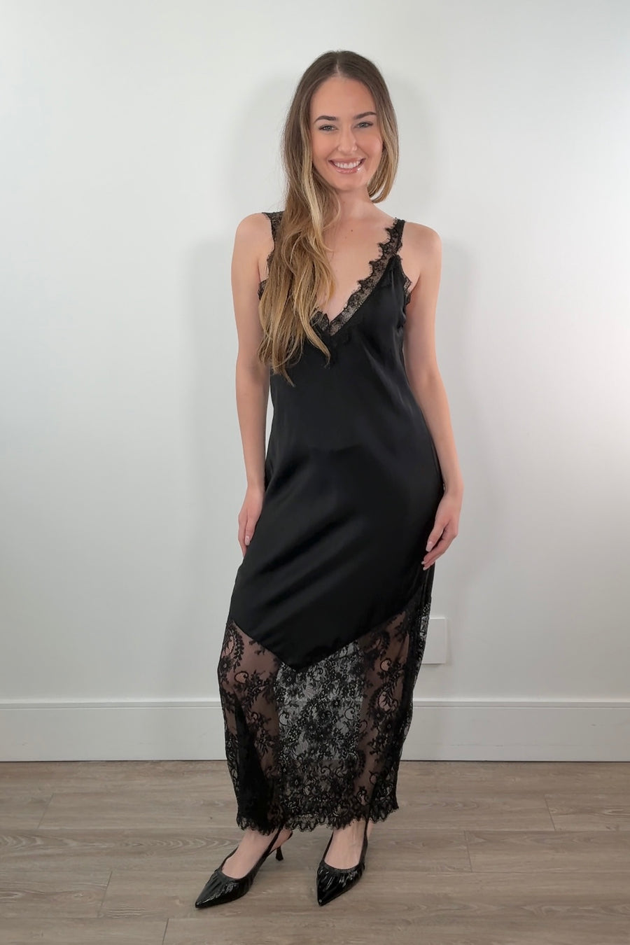 Woman wearing a black satin slip dress with lace details against a white wall.
