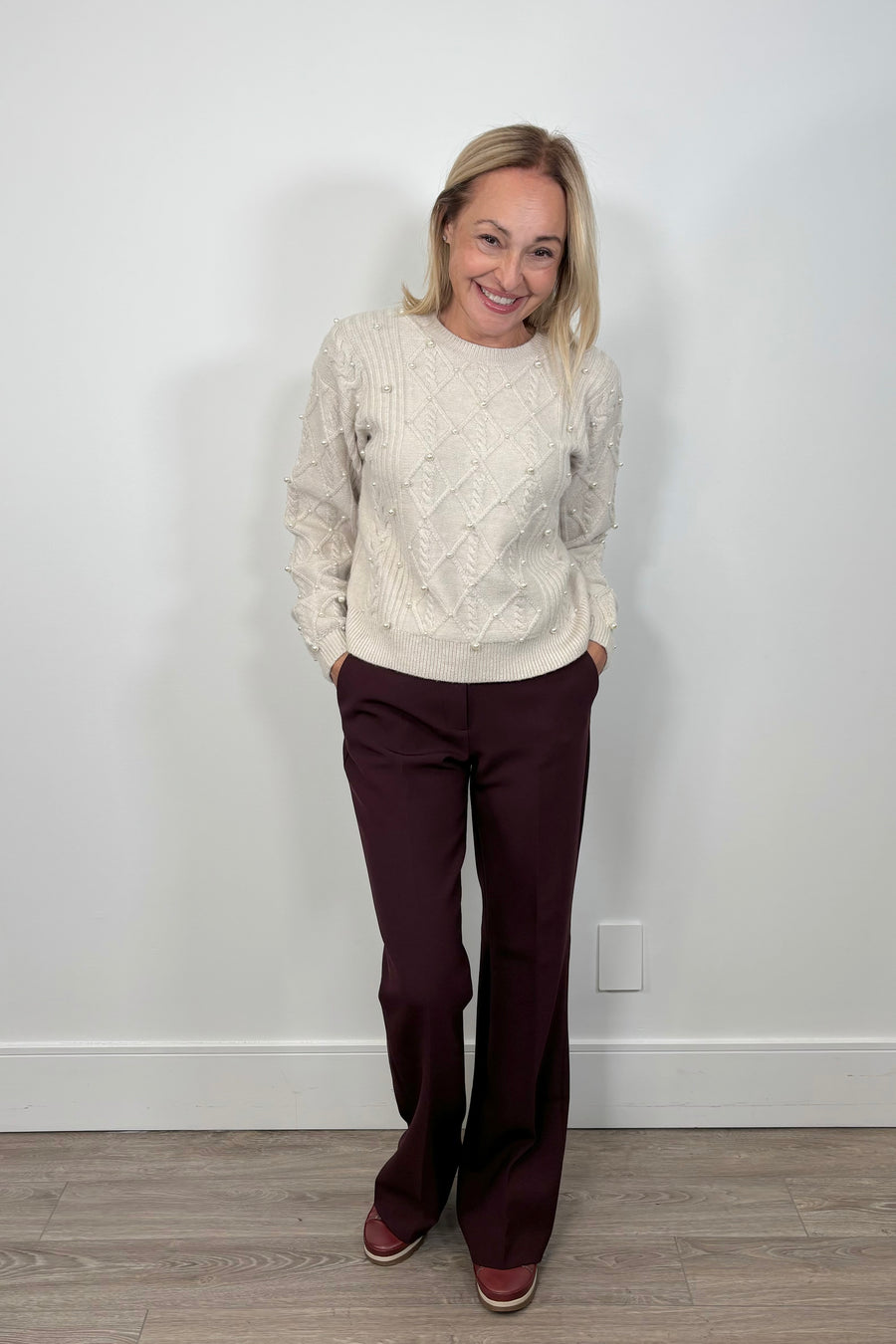 Woman wearing a cream sweater and burgundy pants standing against a white wall.