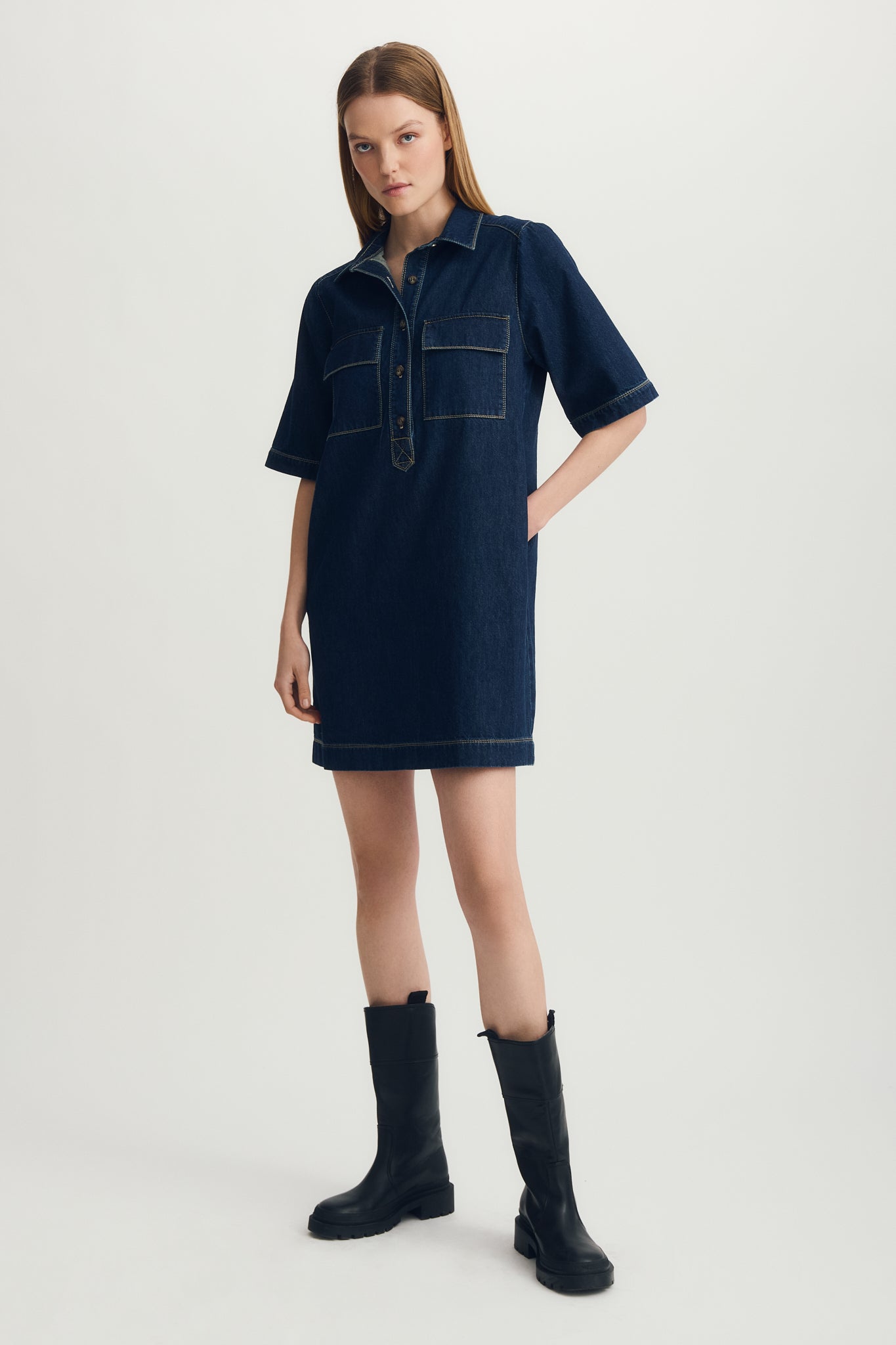 Woman wearing a dark blue denim dress with black boots on a light gray background