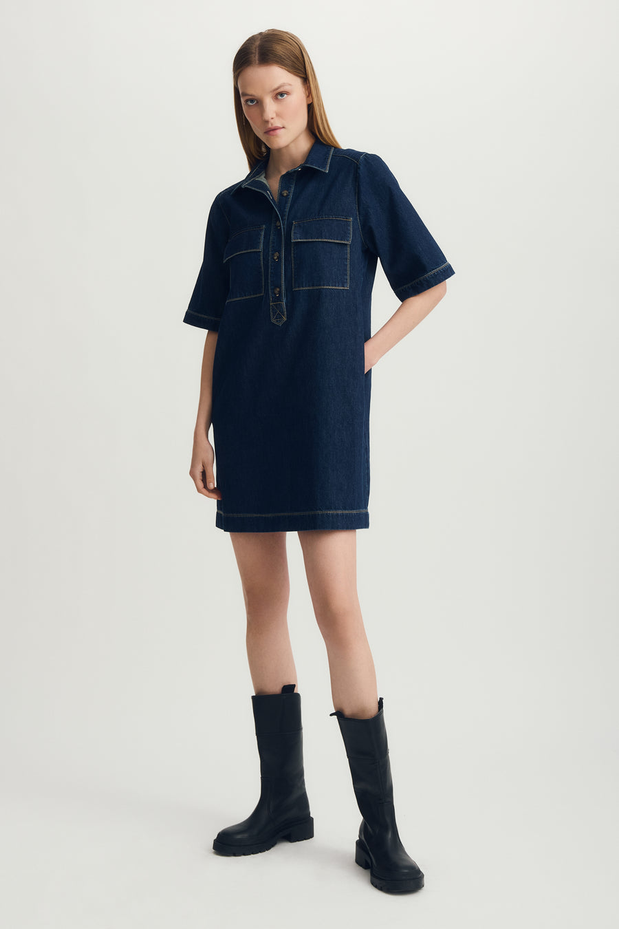 Woman wearing a dark blue denim dress with black boots on a light gray background