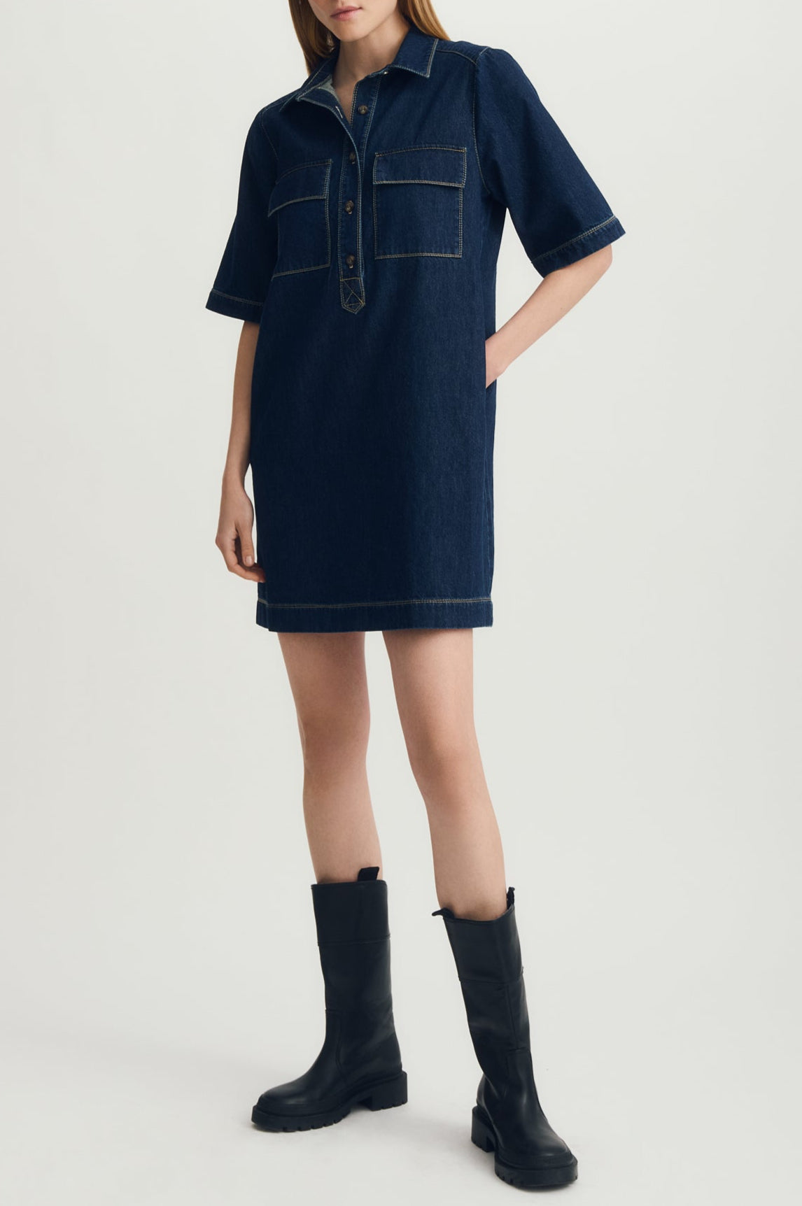 Woman wearing a dark blue denim dress with black boots on a light gray background