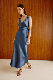 A woman modeling a sleeveless, wrap-style maxi dress in a dusty slate blue satin fabric, standing against a warm wood-paneled wall.