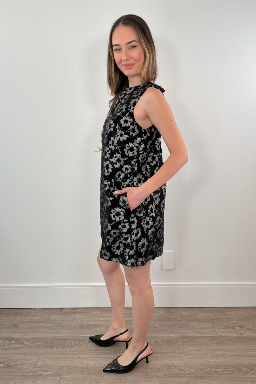 Woman wearing a black floral dress standing against a white wall.