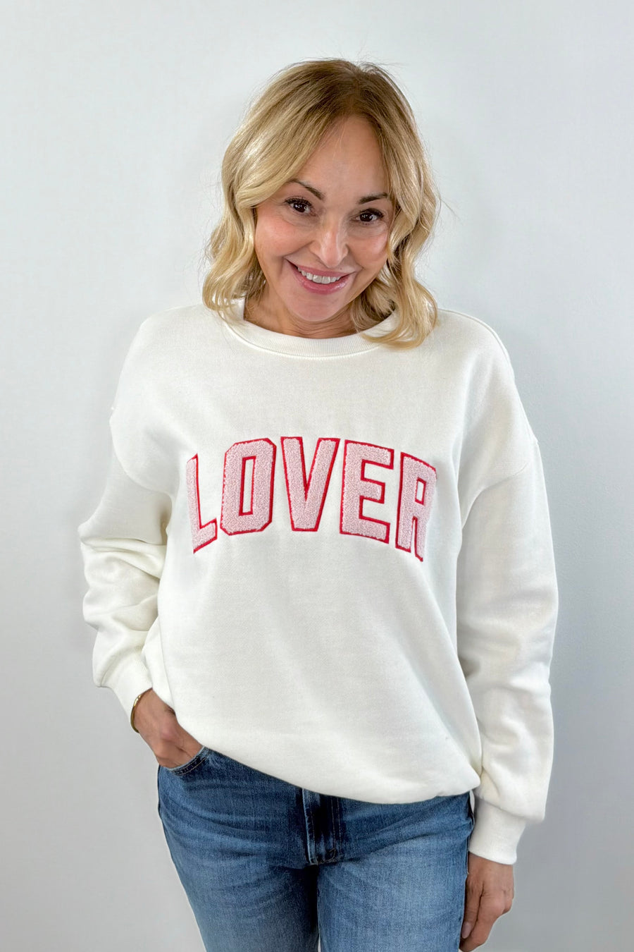 A woman wearing an off white crewneck sweatshirt with the word  “LOVER” written in pink chenille