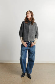 Person wearing a gray and beige sweater with blue jeans against a white wall.