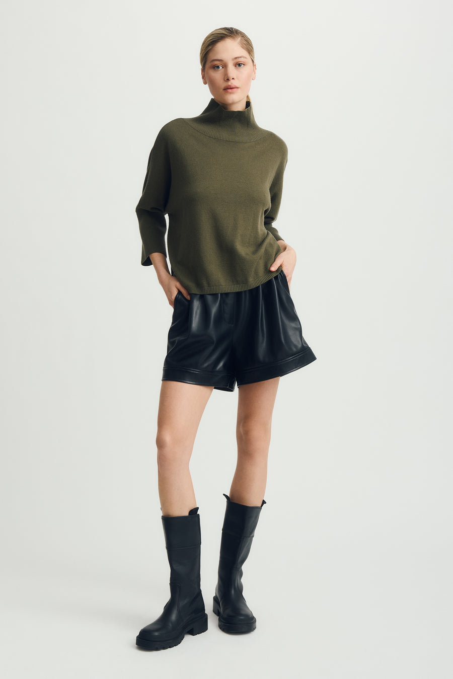 Person wearing a green turtleneck sweater, black leather shorts, and black rain boots on a white background