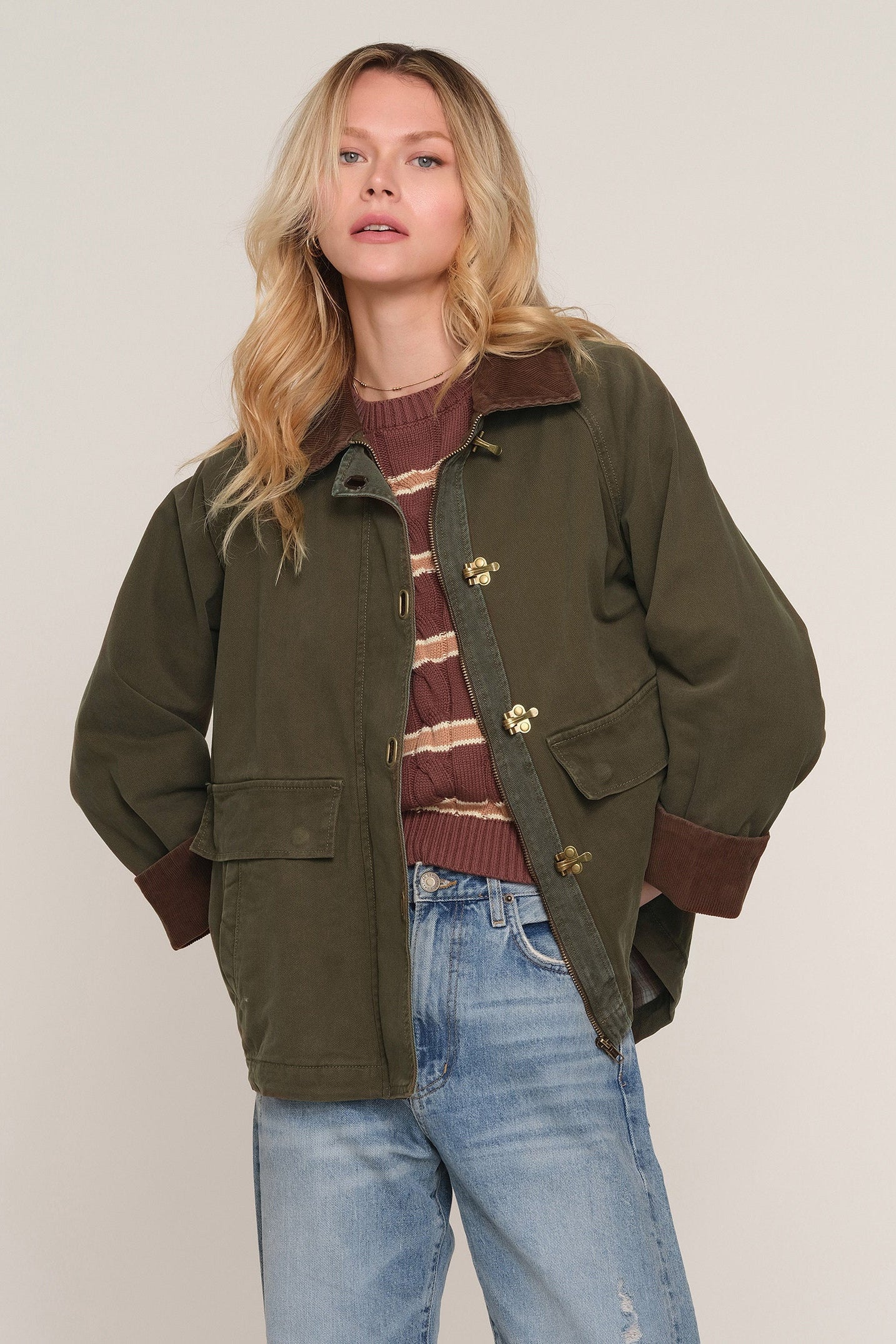 A woman modeling an olive green utility jacket with brown ribbed trim and gold-tone buttons, worn over a brown striped sweater and light blue jeans against a plain light beige background.