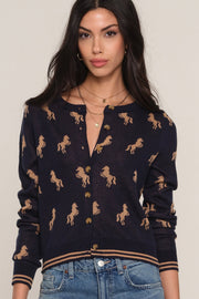 A woman wearing a navy blue knit cardigan with a tan horse pattern, paired with blue jeans, against a plain light beige background.