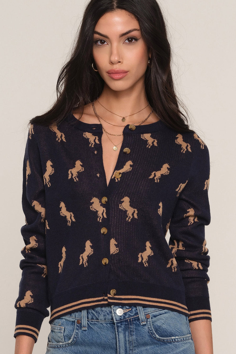 A woman wearing a navy blue knit cardigan with a tan horse pattern, paired with blue jeans, against a plain light beige background.