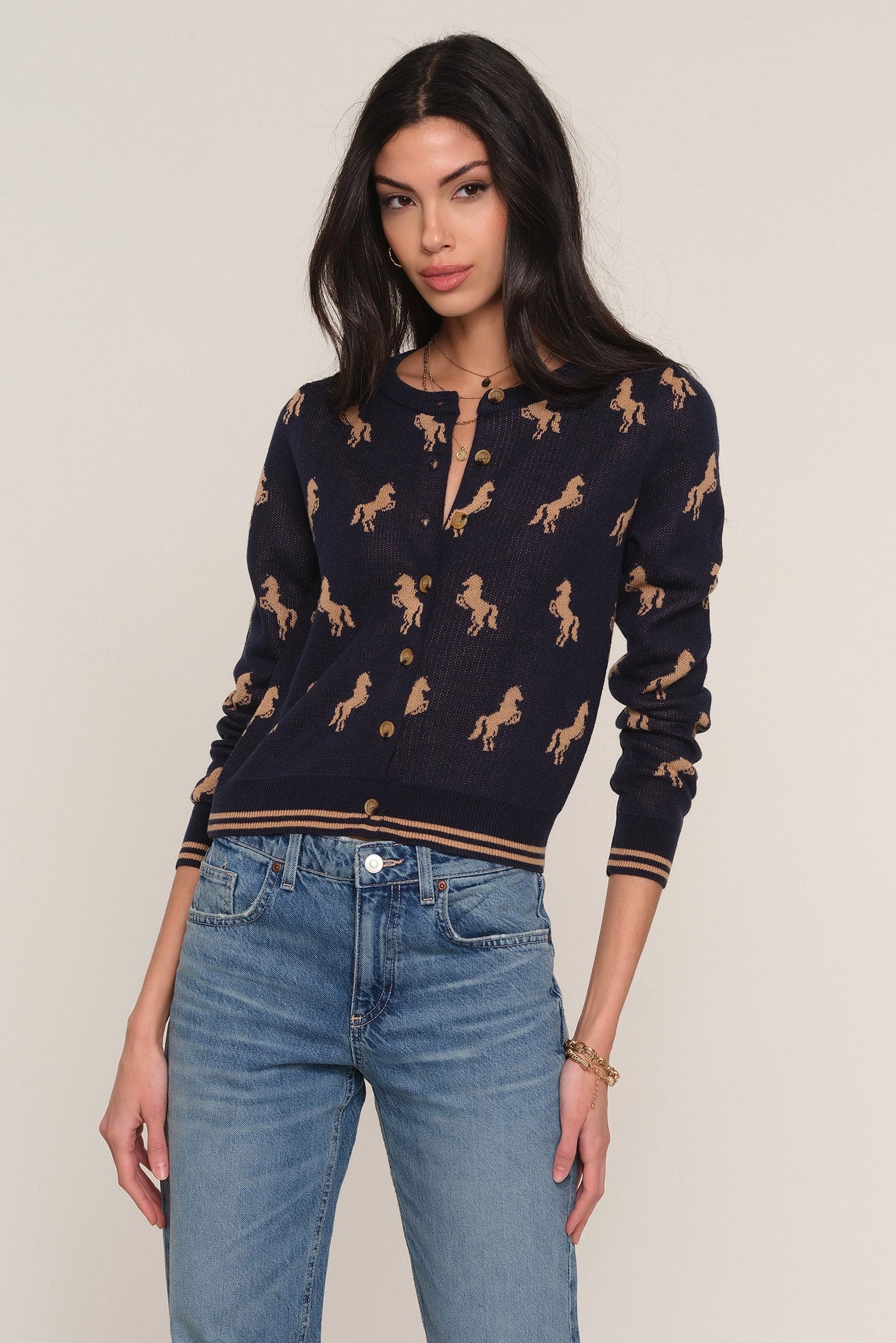 A woman modeling a navy blue knit cardigan with a tan horse pattern, paired with light blue jeans, against a plain beige background.