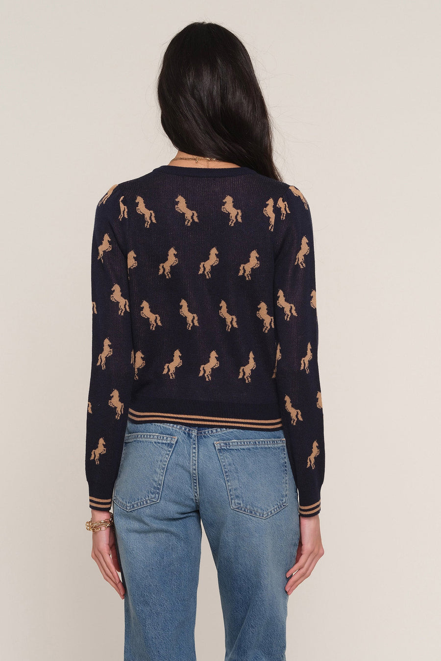 A woman viewed from the back wearing a navy blue long-sleeve sweater with an allover tan horse pattern and light blue jeans, against a plain light beige studio background.