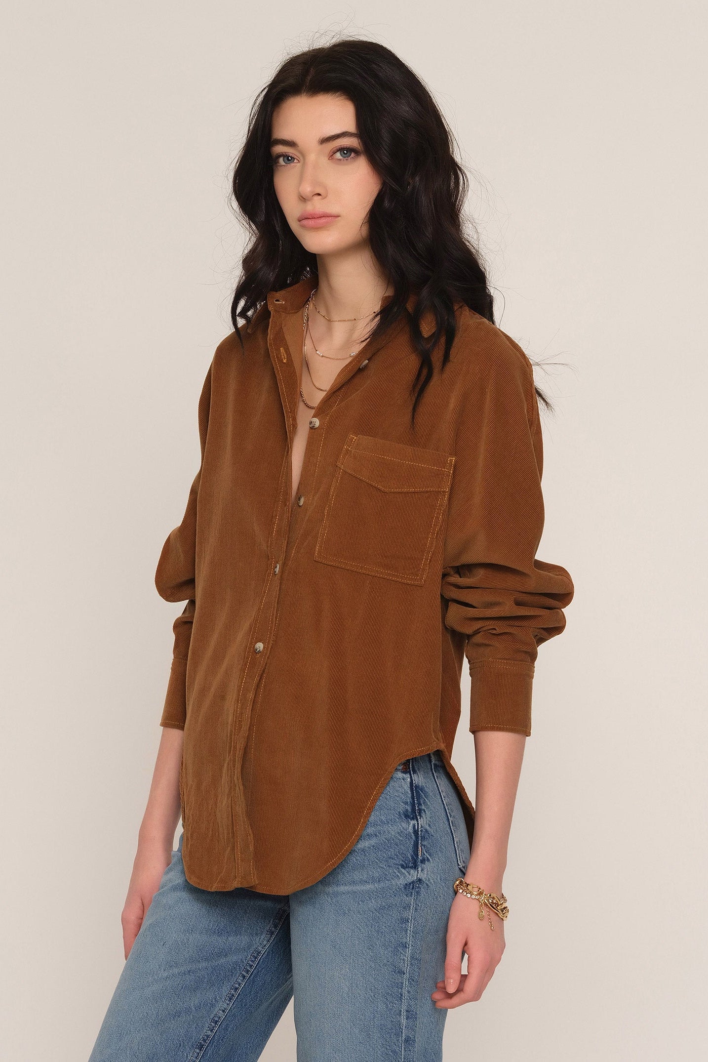 A woman modeling a brown corduroy button-up shirt with a chest pocket, paired with blue jeans, against a plain light beige background.