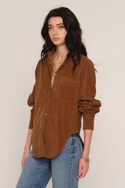 A woman modeling a brown corduroy button-up shirt with a chest pocket, paired with blue jeans, against a plain light beige background.