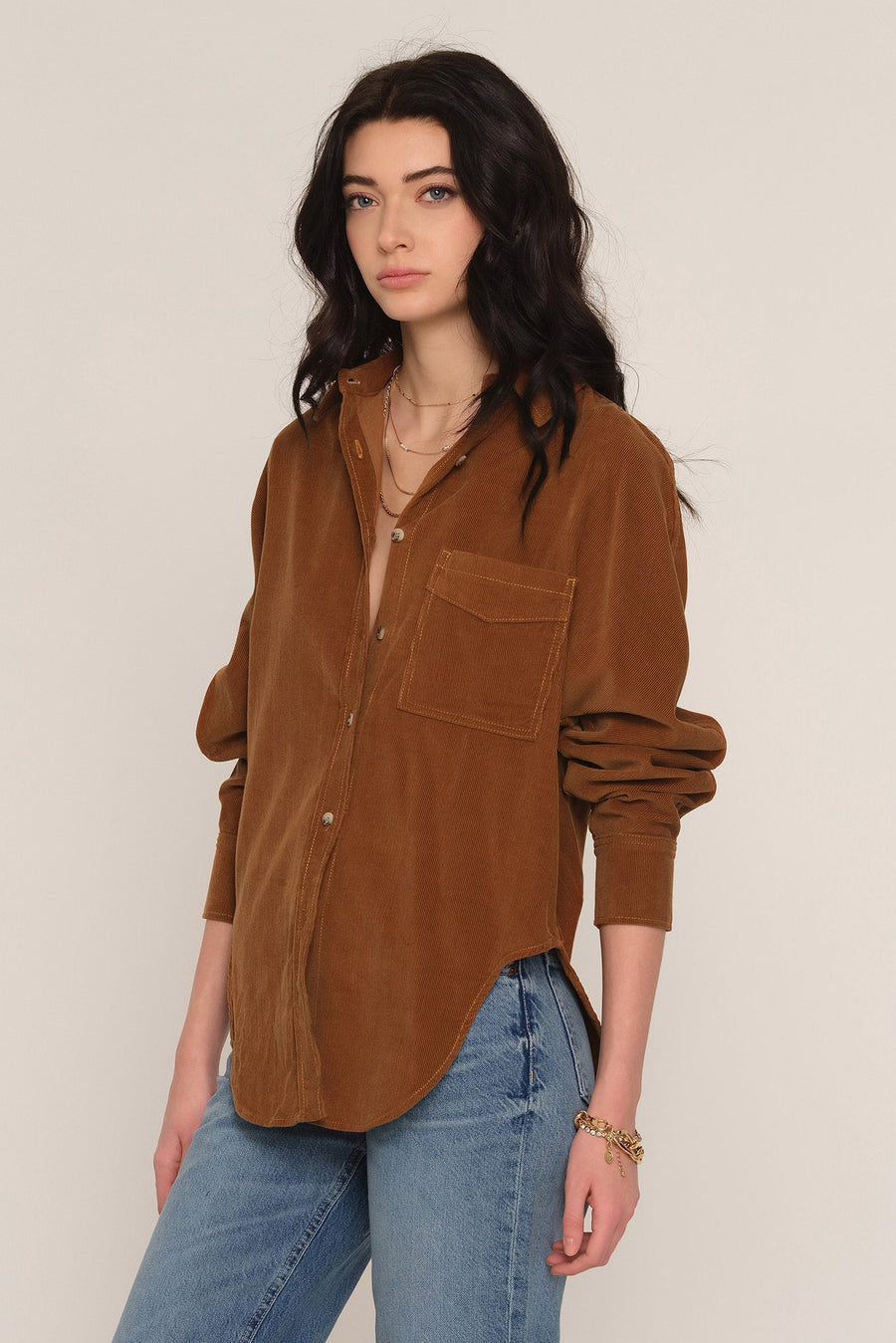 A woman modeling a brown corduroy button-up shirt with a chest pocket, paired with blue jeans, against a plain light beige background.