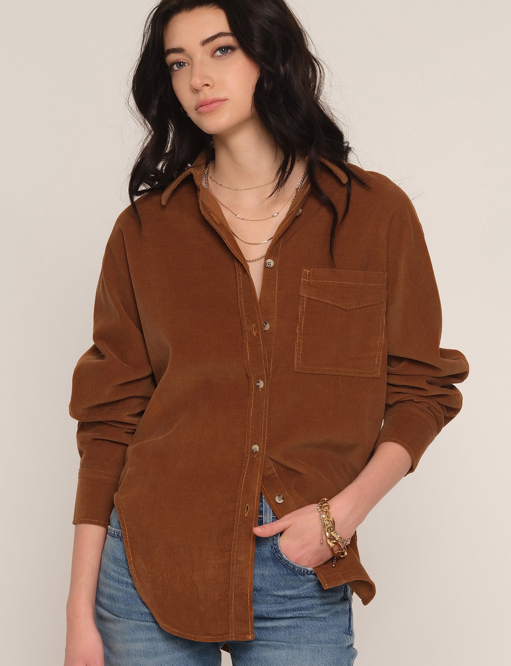 A woman wearing a brown corduroy button-up shirt with a chest pocket, styled with blue jeans and layered gold necklaces, against a plain light beige background.