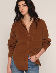 A woman wearing a brown corduroy button-up shirt with a chest pocket, styled with blue jeans and layered gold necklaces, against a plain light beige background.