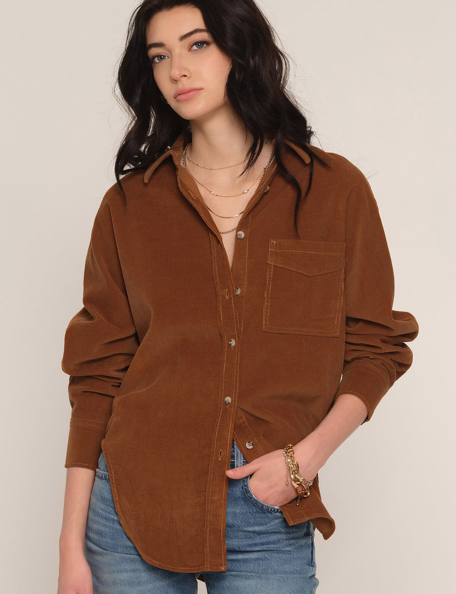 A woman wearing a brown corduroy button-up shirt with a chest pocket, styled with blue jeans and layered gold necklaces, against a plain light beige background.