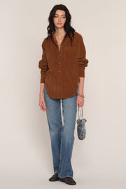 A woman stands against a plain light beige studio background wearing an oversized brown corduroy button-up shirt
