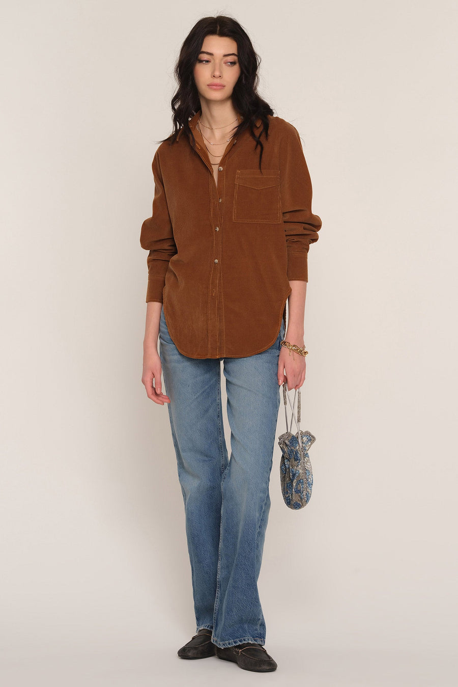 A woman stands against a plain light beige studio background wearing an oversized brown corduroy button-up shirt