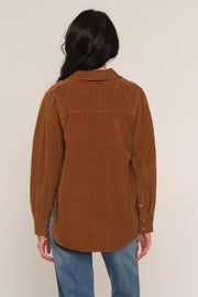 A woman viewed from the back wearing a brown corduroy button-up shirt jacket and blue jeans against a plain light beige studio background.