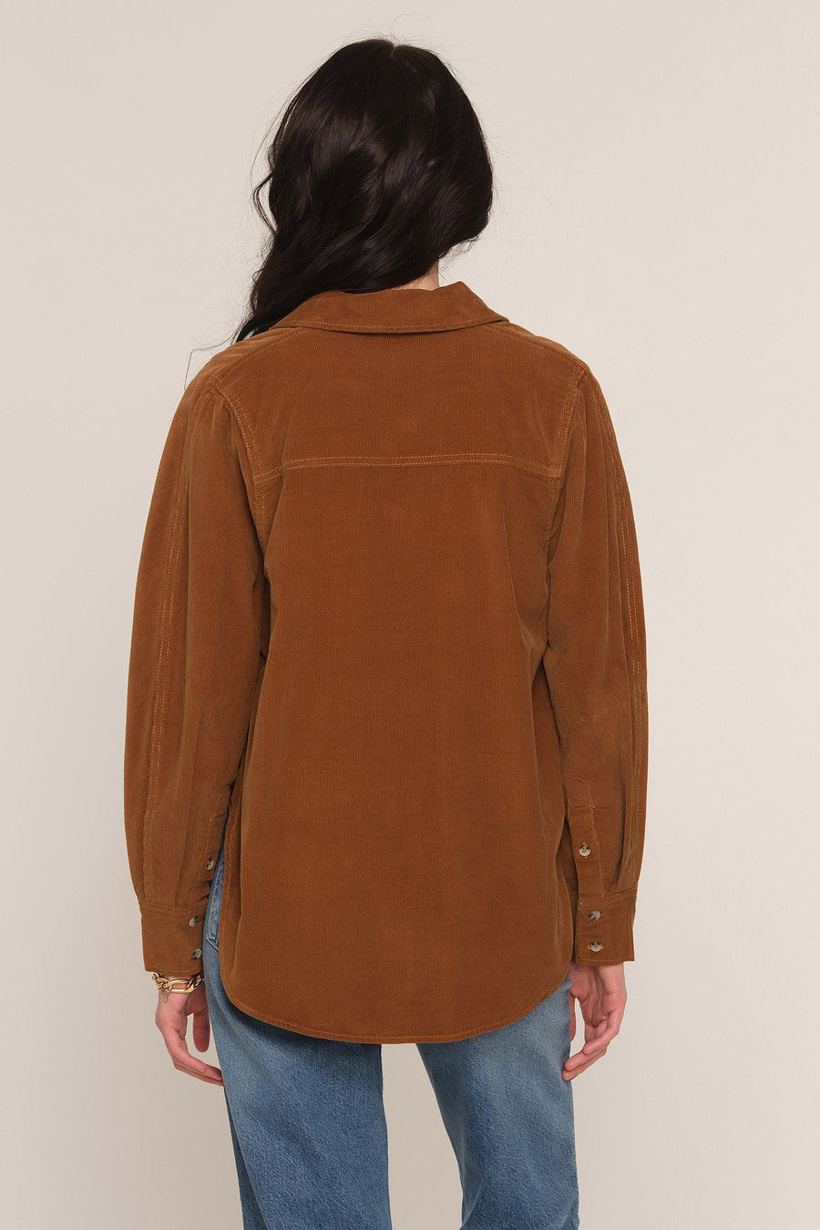 A woman viewed from the back wearing a brown corduroy button-up shirt jacket and blue jeans against a plain light beige studio background.