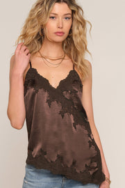 A woman wearing a brown satin camisole top with black lace trim and a distressed, tattered hem, paired with blue jeans against a light beige background.