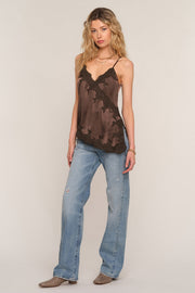 A woman modeling a brown sheer lace camisole top with black trim, paired with light-wash blue jeans and brown boots, against a plain beige studio background.