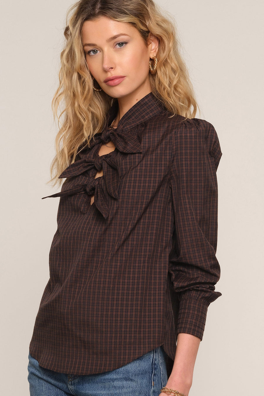 A woman wearing a dark brown plaid long-sleeve blouse with a large ruffled bow at the collar and keyhole cutout,