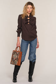 A model stands against a light beige studio background wearing a dark brown ruffled long-sleeve blouse