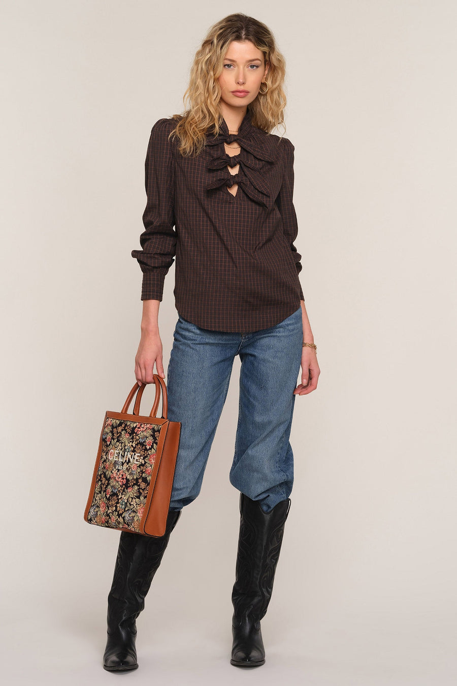 A model stands against a light beige studio background wearing a dark brown ruffled long-sleeve blouse