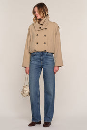 A model is shown from the head to toe wearing a cropped beige double-breasted trench jacket, blue jeans, and brown shoes, holding a small beige chain bag against a plain light beige studio background.