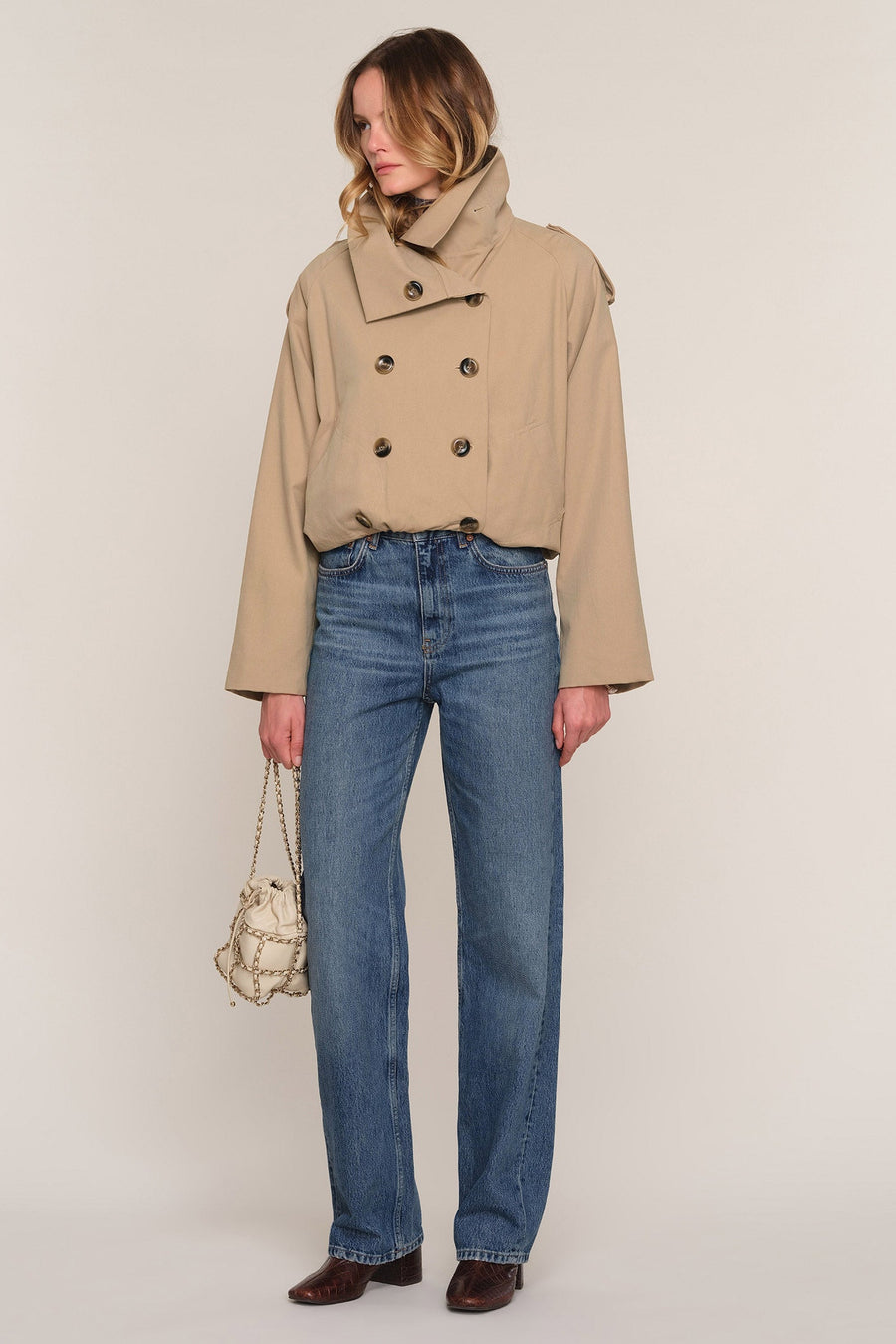 A model is shown from the head to toe wearing a cropped beige double-breasted trench jacket, blue jeans, and brown shoes, holding a small beige chain bag against a plain light beige studio background.