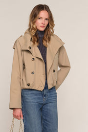 A woman modeling a cropped beige double-breasted jacket over a dark blue plaid turtleneck sweater and blue jeans, standing against a plain light beige studio background.
