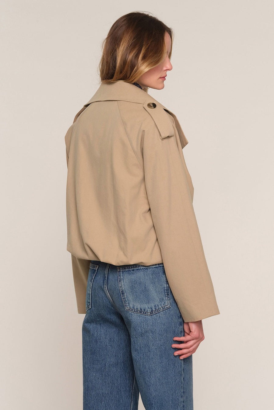A woman shown from the back wearing a cropped beige trench jacket and blue jeans against a plain light beige studio background.